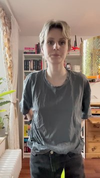 Siva majica z ohlapnim krojem in zvitim rokavom, v kombinaciji s črnimi hlačami. Notranji prostor z regali in rastlino v ozadju.