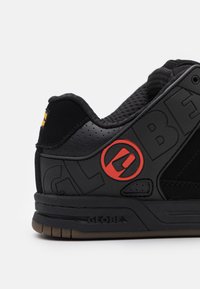 Zapatilla negra con una parte superior texturizada, que presenta un logo rojo prominente y detalles perforados. Suela de goma con un sutil patrón de banda.
