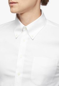 Close-up van een persoon in een wit overhemd met knopen, een button-down kraag en een borstzak aan de linkerkant.