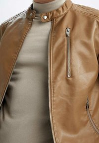 Primo piano di una giacca di pelle marrone chiaro con cerniere argentate indossata sopra un maglione a collo alto beige.