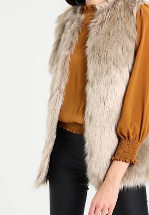 Personne portant une veste en fausse fourrure beige sur un chemisier moutarde à manches longues avec poignets smockés et un pantalon noir, debout devant un fond uni.