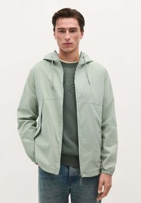 Lys grøn hættejakke lavet af glat stof, med lynlås foran, elastiske manchetter og et simpelt design, båret over en mørk sweatshirt.