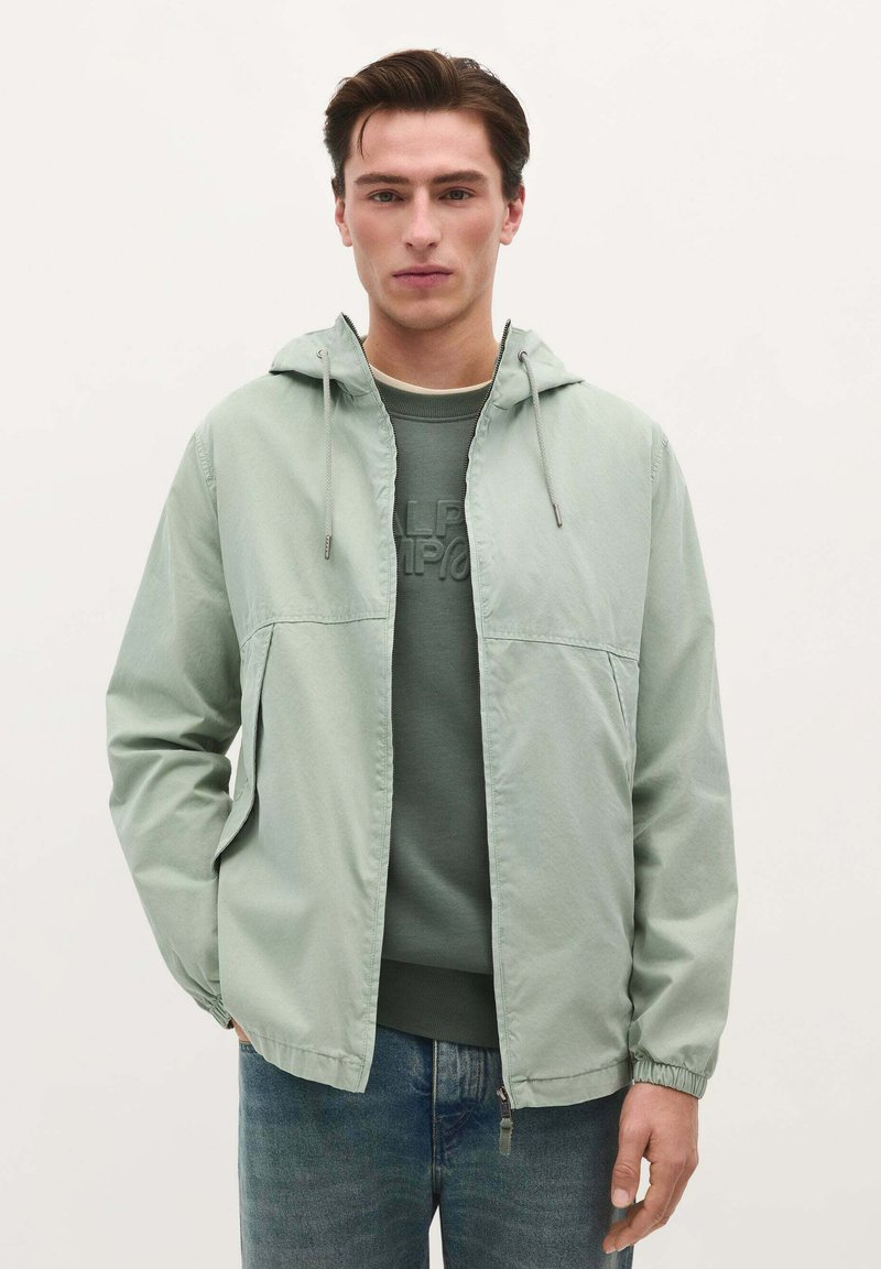 Lys grøn hættejakke lavet af glat stof, med lynlås foran, elastiske manchetter og et simpelt design, båret over en mørk sweatshirt.