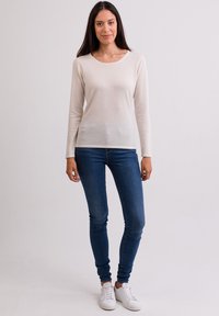 Helles crèmefarbenes Langarmshirt mit rundem Ausschnitt, kombiniert mit dunkelblauen Skinny-Jeans und weißen Sneakers. Glatte Stricktextur.