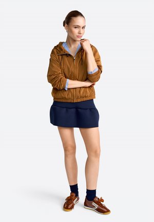 Frau steht mit verschränkten Armen, trägt eine braune Jacke, einen marineblauen Rock, dunkle Socken und braune Turnschuhe vor einem schlichten weißen Hintergrund.