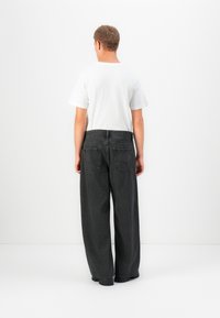 Mann som står med ryggen til, iført en hvit T-skjorte og løse sorte denimjeans med kraftige svarte sko mot en ensfarget hvit bakgrunn.