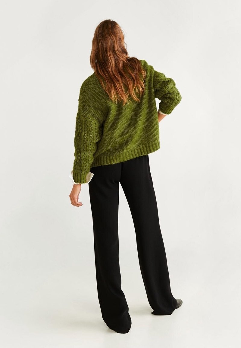 Femme aux longs cheveux bruns portant un pull vert à grosse maille et un pantalon noir à jambes larges, debout de dos à la caméra.