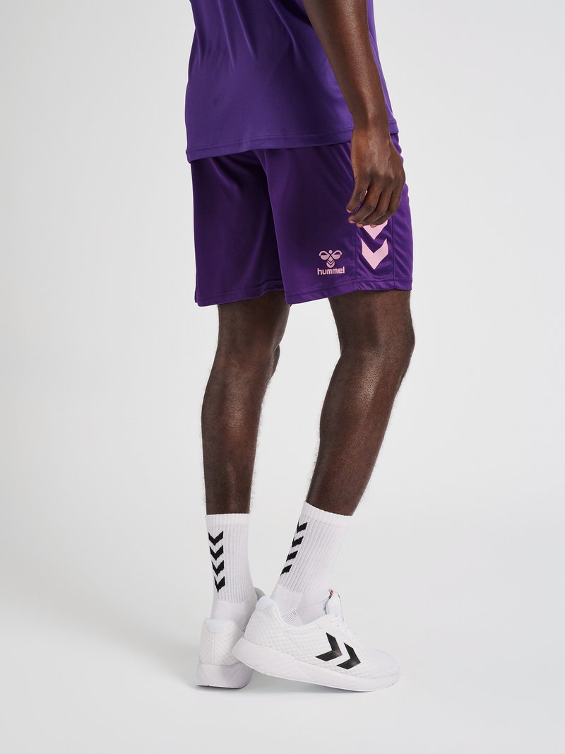 Person iført lilla sportsshorts, hvide sokker med sorte chevrons og hvide sneakers med sorte chevron-logoer på en ensfarvet baggrund.