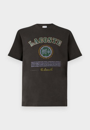 Juodas medvilnės marškinėliai su apvaliu žaliu ir geltonu piešiniu, kuriame bold rašmenimis užrašyta "LACOSTE" ir "TENIS HERITAGE".