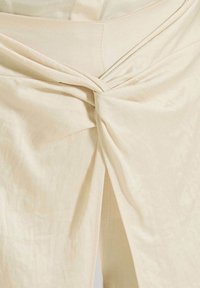 Beige stof met een gedraaide knoopdetail bij de taille, zachte textuur en een vloeiende val. Kenmerkt zich door nette stiksels en een subtiele glans.