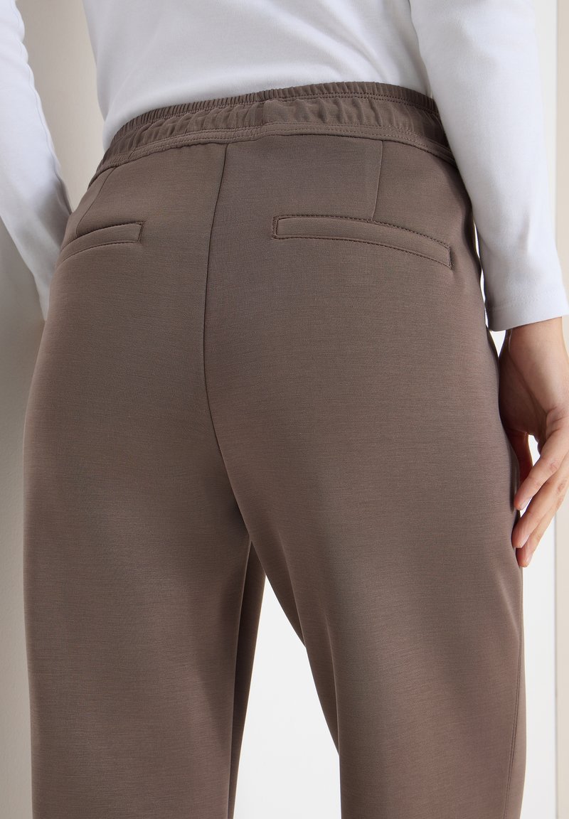 Vue arrière d'une personne portant un pantalon taupe à taille élastique et un haut blanc à manches longues, main reposant sur le côté, fond neutre.