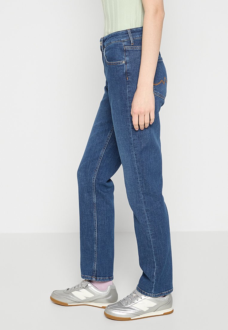 Blaue Jeans im geraden Schnitt mit orangen Nähten an den Gesäßtaschen, kombiniert mit silbernen Sportschuhen.