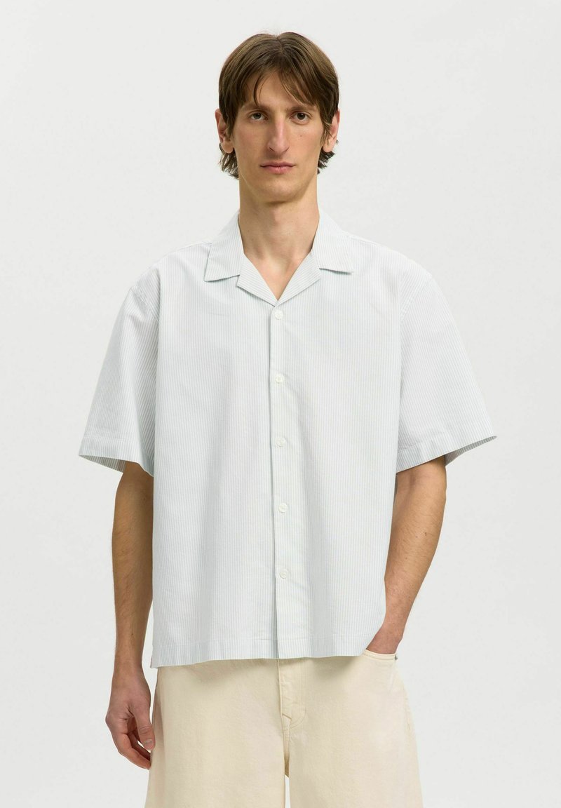 Modèle masculin portant une chemise bleu clair à manches courtes boutonnée et un pantalon beige, debout sur un fond blanc uni.