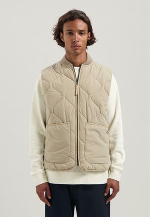 Beige gewatteerde gilet met een geribbelde kraag, een rits aan de voorkant en twee grote zakken. Draagt over een witte trui, heeft een gladde textuur.