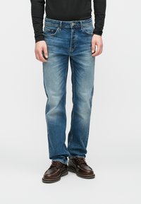 Τζιν παντελόνι σε μέτρια μπλε απόχρωση με ξεθωριασμένη υφή, ίσια γραμμή, πέντε τσέπες και κλείσιμο με κουμπί, συνδυασμένο με καφέ δερμάτινα παπούτσια με κορδόνια.