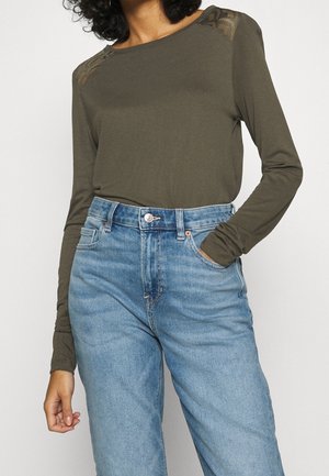 Femme portant un haut à manches longues vert olive avec des détails en dentelle aux épaules et un jean bleu clair taille haute, une main dans la poche.