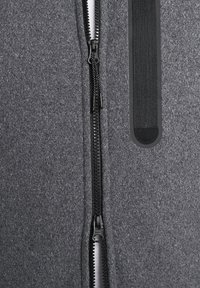 Šedý plstěný materiál s černým středovým zipem a černým elastickým detailem. Texturovaný povrch, minimalistický design, funkční kovové prvky.