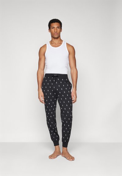 Pantalons de survêtement noirs avec un motif de logo blanc, taille élastique et poignets. Associés à un débardeur blanc uni. Le mannequin est pieds nus.