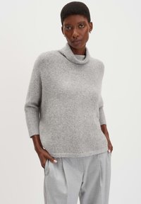 Hellgrauer Rollkragenpullover aus weichem Strickmaterial, mit Dreiviertelärmeln und lockerer Passform, kombiniert mit lockeren grauen Hosen.
