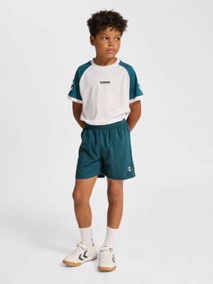 Junge steht mit den Händen hinter dem Rücken, trägt weiße und türkisfarbene Sportbekleidung, weiße Socken und weiße Sneakers vor einfarbigem Hintergrund.