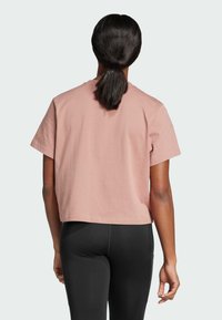 Zkrácené tričko s krátkým rukávem v světle růžové barvě z bavlny, s jednoduchým designem. Zadní pohled ukazuje hladkou texturu a uvolněný střih. Viditelné černé legíny.