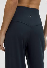 Krupan pogled na leđa osobe koja nosi tamne sportske hlače visokog struka s logom. Ruka je položena blizu struka.