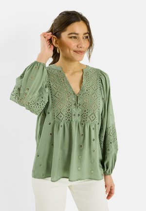 Mujer con blusa de manga larga verde claro con patrones calados bordados, combinada con pantalones blancos, sonriendo y tocándose el cabello.