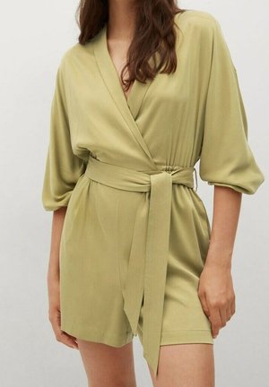 Woman wearing a light olive green wrap dress with three-quarter sleeves and a tied waist belt, standing against a plain background.