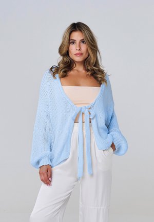Femme aux cheveux ondulés portant un cardigan tricoté bleu clair noué à l'avant sur un top beige tube et un pantalon blanc ample, debout sur un fond gris.