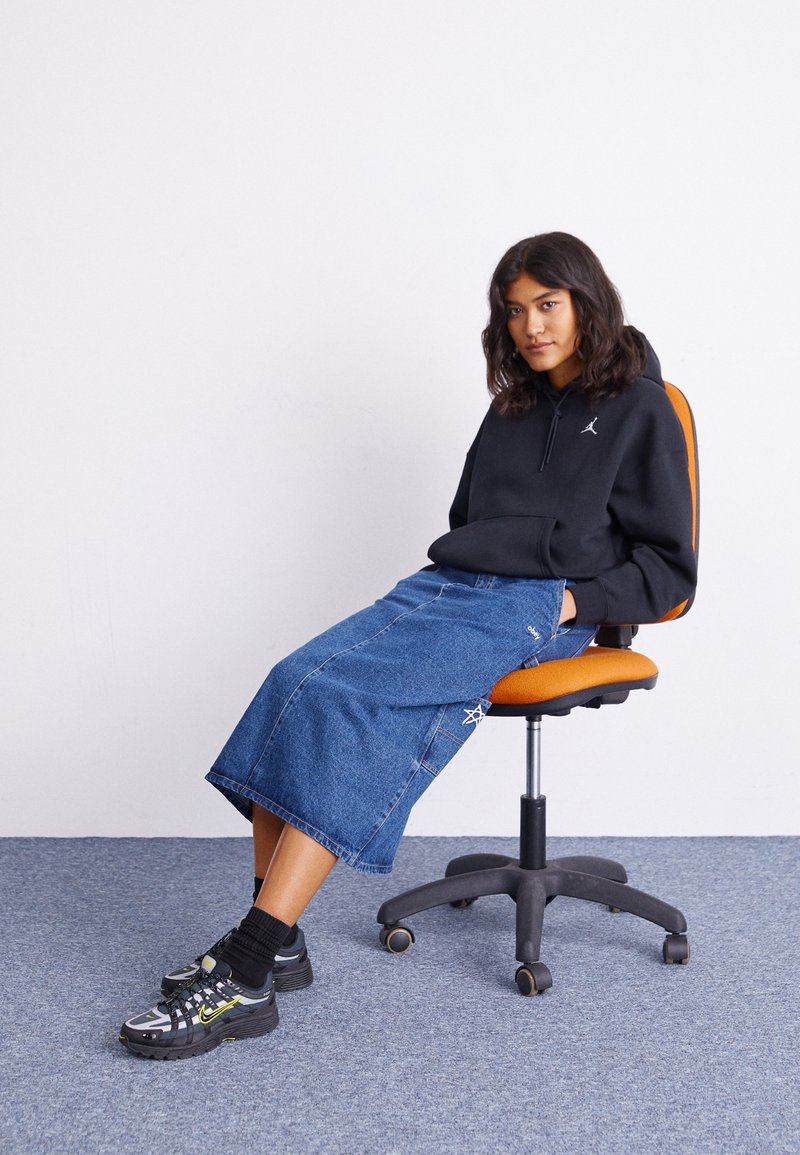 Sudadera con capucha negra con logo, falda midi de mezclilla con detalles de costura y zapatillas negras con acentos amarillos, sentado en una silla naranja.