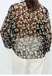 Blouse noire transparente avec imprimé floral marguerite. Comprend des manches longues, une coupe décontractée et une texture semi-transparente.