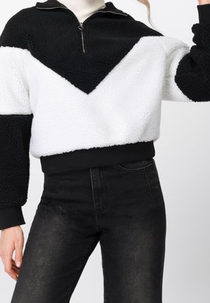 Personne portant un pull pelucheux noir et blanc avec une fermeture éclair au quart et un jean noir taille haute sur fond blanc.