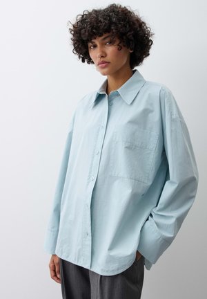 Femme aux cheveux bouclés portant une chemise ample bleu clair boutonnée et un pantalon gris, debout devant un fond clair uni.
