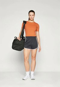 Oranž fiteeritud lühikeste varrukatega topp, mustad spordilühikesed püksid valge äärisega ja valged tossud. Kandes musta spordikotti rihmaga.