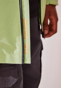 Hellgrüne wasserdichte Jacke mit einem schwarzen Reißverschluss, verfügt über orangefarbene Textdetails und eine glatte Textur. Akzentuiert mit einem markanten Design.