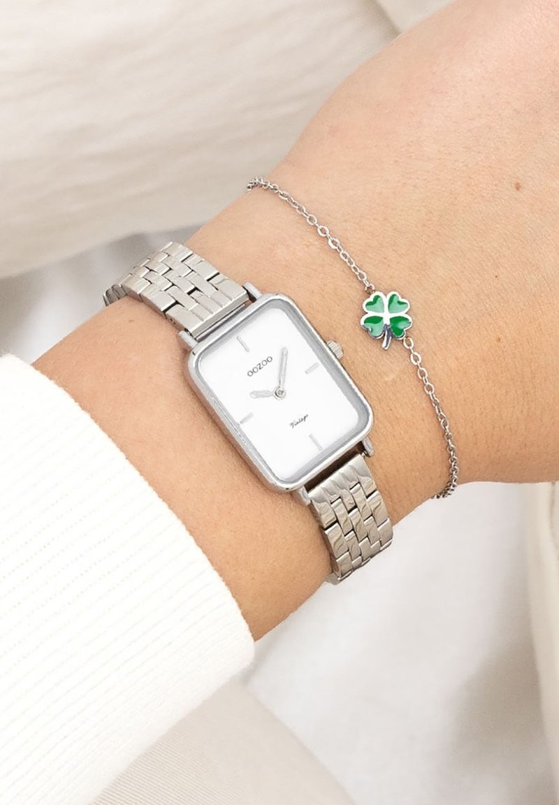 Montre-bracelet en métal argenté avec un cadran rectangulaire, un fond blanc et deux aiguilles. Portée avec un bracelet en chaîne argenté orné d'un charme en forme de trèfle à quatre feuilles vert.