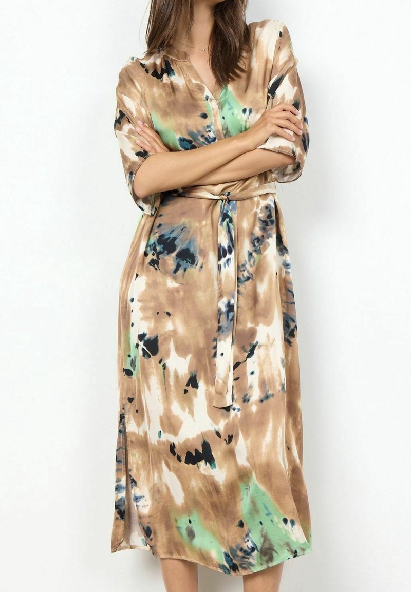 Femme portant une robe tie-dye courte à manches courtes avec une ceinture à la taille, aux couleurs beige, vert, noir et blanc, debout les bras croisés.