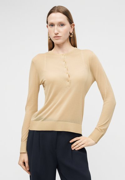 Γυναίκα που φοράει μπεζ μακρυμάνικο πουκάμισο henley με κουμπιά και μπλε σκούρο παντελόνι φαρδιάς γραμμής, στέκεται με το ένα χέρι στη μέση μπροστά σε απλό φόντο.