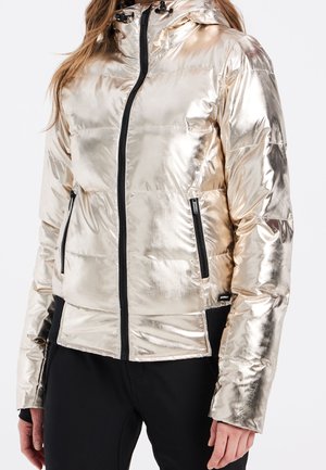 Kobieta w błyszczącej metalicznej złotej kurtce puchowej z czarnymi zamkami i czarnych spodniach, stojąca na białym tle.