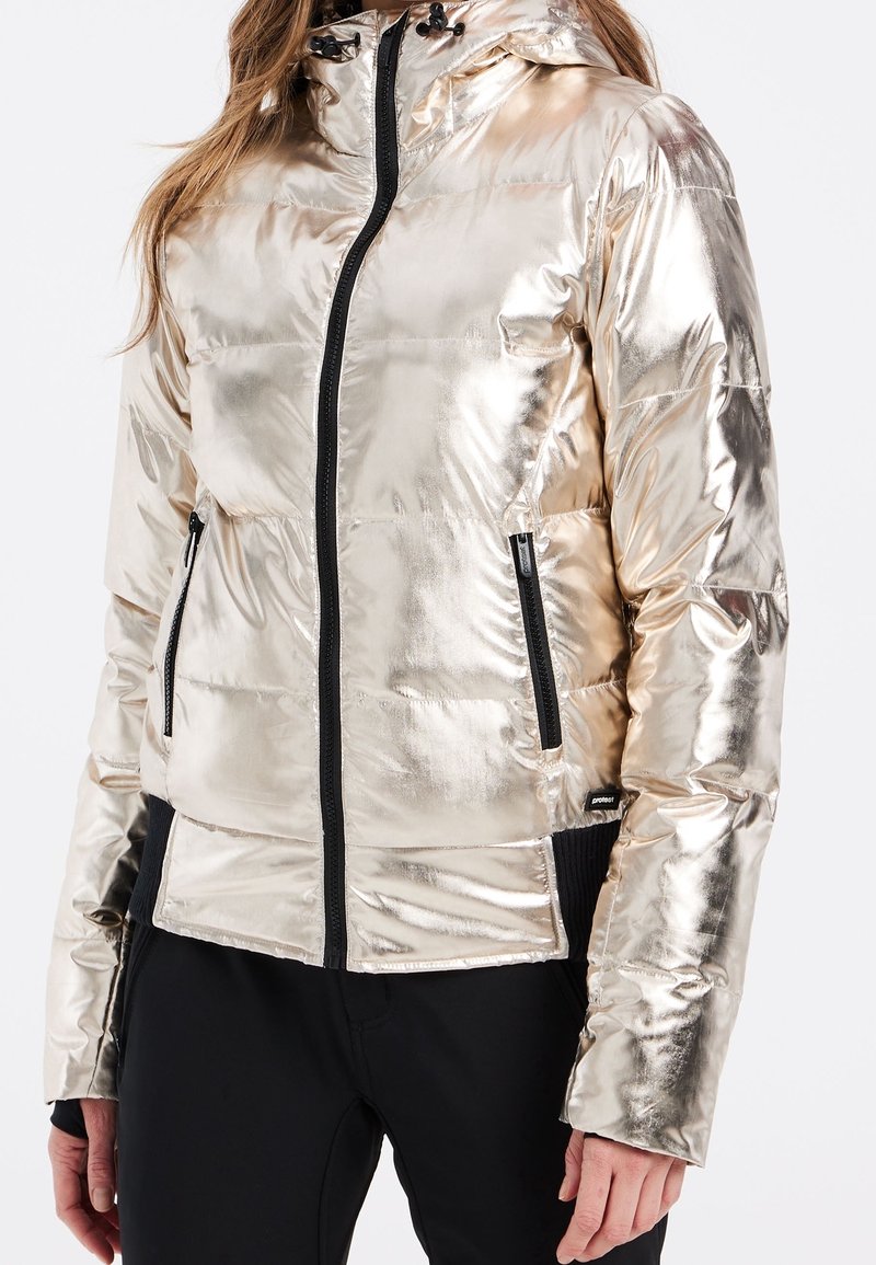 Femme portant une veste matelassée métallique dorée brillante avec des fermetures éclair noires et un pantalon noir, debout sur fond blanc.
