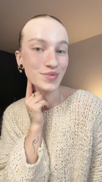 Svetlý pletený sveter s textúrovaným vzorom, zlaté kruhové náušnice a niekoľko prsteňov. Viditeľné tetovanie na ruke. Mäkké osvetlenie v pozadí.