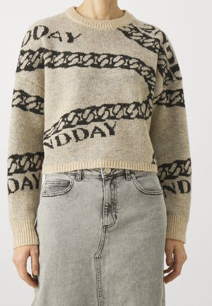 Person iført beige strikket sweater med sort kædemønster og teksten "NO DAY", kombineret med en lysegrå denim nederdel.