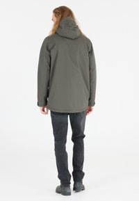 Giacca da esterno verde con cappuccio, caratterizzata da un tessuto testurizzato e una vestibilità rilassata, abbinata a jeans grigio scuro e stivali robusti.