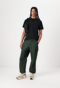 Svart bomulls t-shirt med lila logotyp, gröna cargobyxor med fickor och beigea sportskor. Inkluderar en liten svart axelväska.