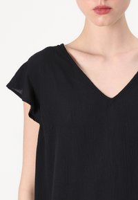 Svart kortermet topp med V-hals, laget av teksturert stoff. Har en løs passform og en enkelt side pocket.