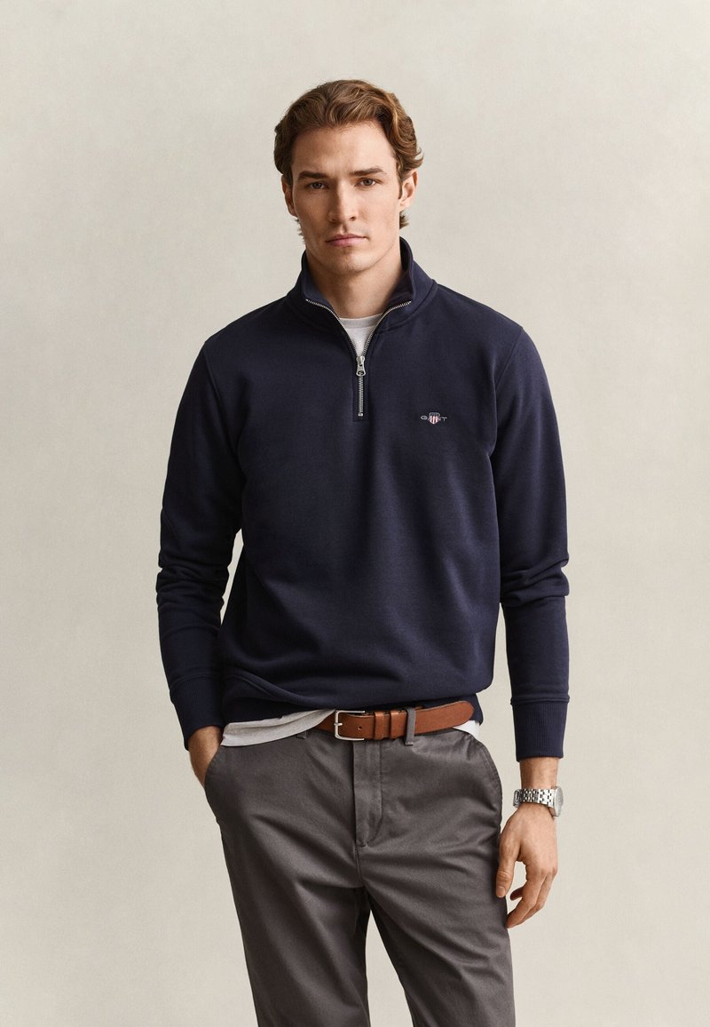 Junger Mann trägt einen marineblauen Pullover mit Viertelreißverschluss, graue Hose, braunen Gürtel und eine silberne Armbanduhr, steht mit einer Hand in der Tasche.