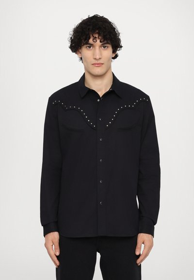 Chemise noire à boutons avec manches longues, présentant un design de suture courbée et des clous métalliques le long des épaules. Deux poches poitrine incluses.