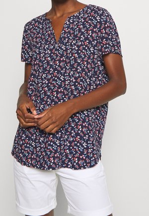 Dunkelhäutige Person trägt eine marineblaue kurzärmelige Bluse mit Blumenmuster und weiße hochgekrempelte Shorts, die Hände vor der Taille gefaltet.