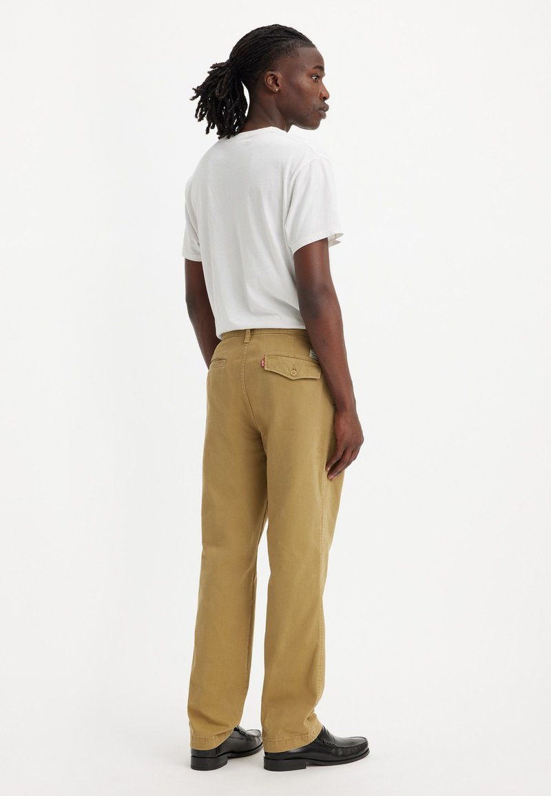 Pantalones de algodón beige con corte recto, bolsillos traseros y costuras en contraste. Llevados con una camiseta blanca sencilla y zapatos negros.