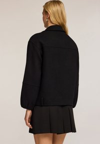 Zwarte wolmix jas met een kraagachtige halslijn, verlaagde schouders en brede mouwen. Voorzien van een ceintuur in de taille en achtersteemdetails.